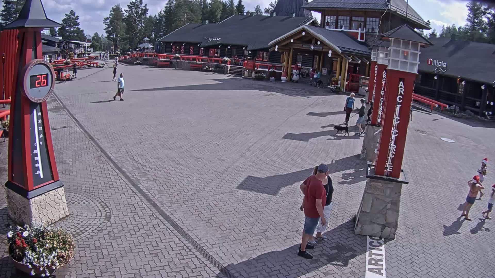 A paved plaza with several people walking around in front of a building on a sunny day,  near tall red posts with white lettering.