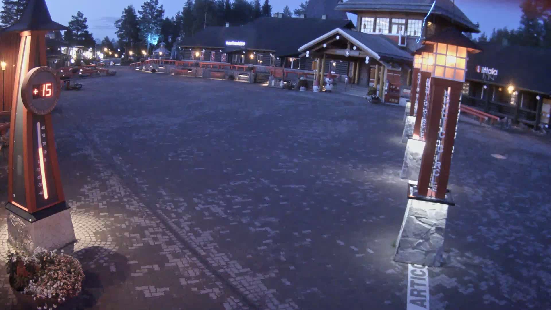 A paved plaza at dusk, with several illuminated buildings and decorative light posts, shows a temperature of 15 degrees.