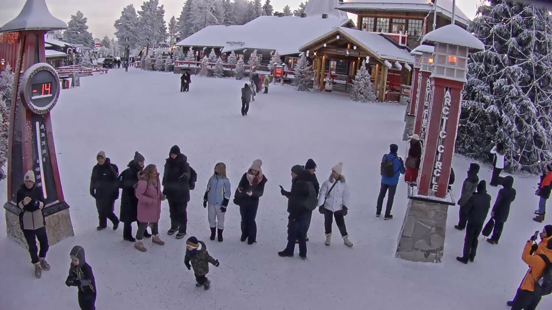 People in winter clothing stand in a snow-covered outdoor area marked by "ARCTIC CIRCLE" pillars and a large thermometer displaying -14 degrees, with festive buildings and frosted trees under an overcast winter sky.
