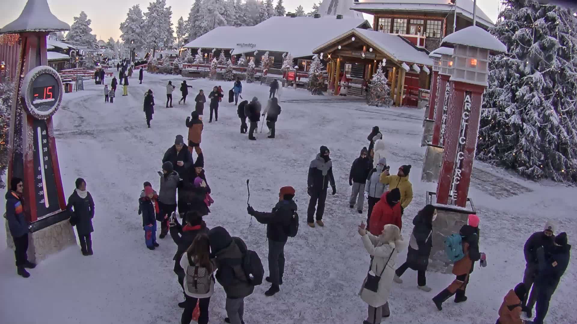 Numerous people in heavy winter clothing are seen walking and gathering on a snow-covered outdoor square, flanked by snow-dusted buildings and trees, with a large thermometer displaying -15 degrees and "Arctic Circle" signs, all under an overcast, cold winter sky.