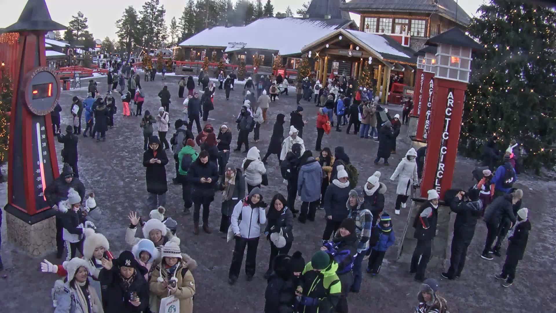 A large crowd of people, bundled in heavy winter clothing, gathers outdoors in a snowy village square on an overcast winter day.