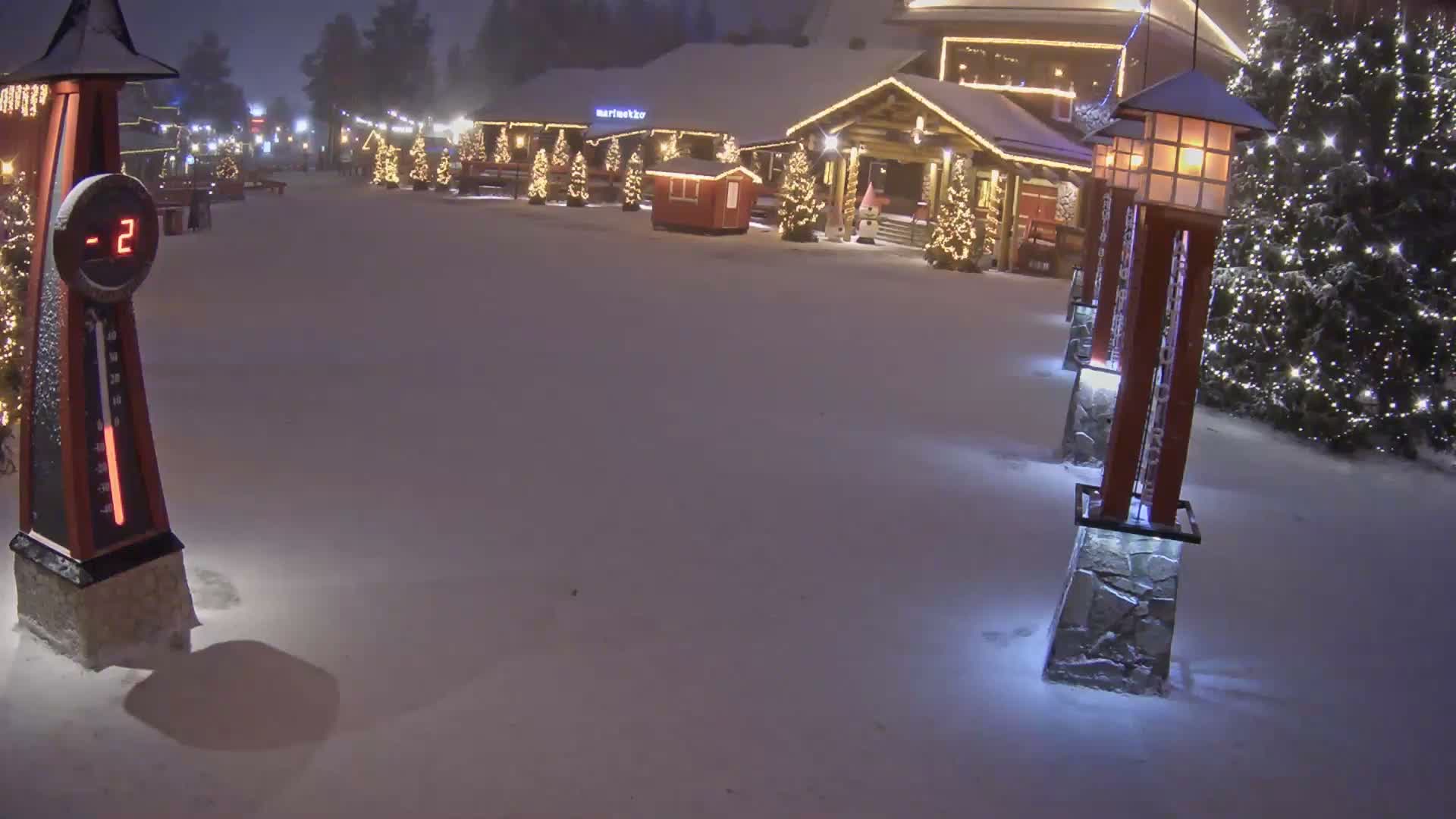 The image displays a snow-covered outdoor village square on a clear, cold night, illuminated by numerous festive holiday lights on buildings and decorated trees, with a prominent thermometer indicating a temperature of -2 degrees Celsius.