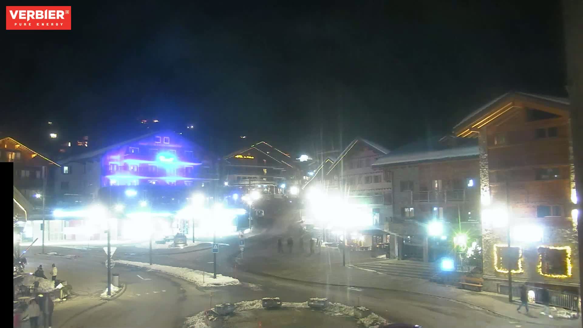 A nighttime scene of a snow-covered town square with brightly lit buildings and a few pedestrians.