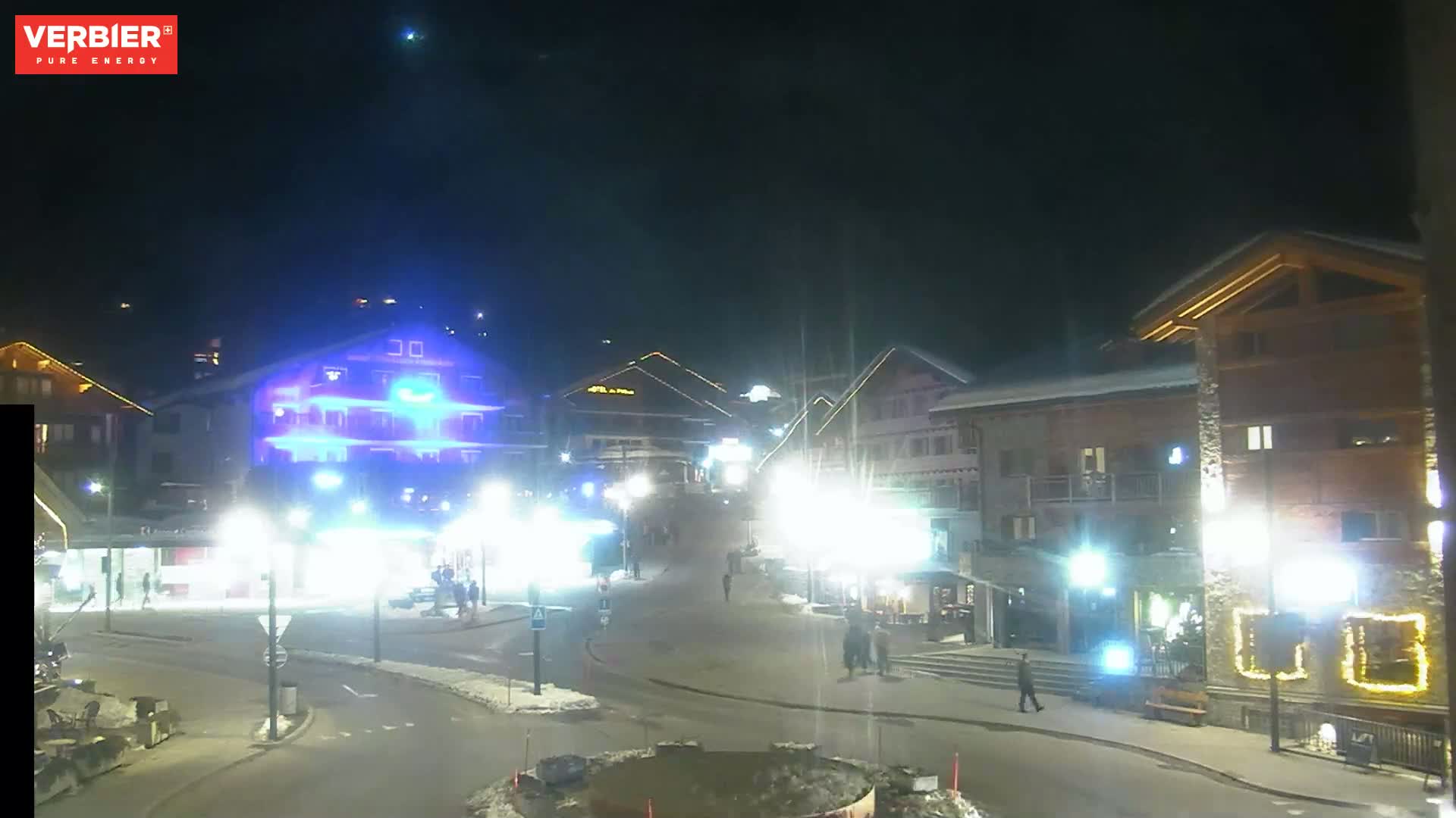 A nighttime, snow-covered town square is illuminated by streetlights, showing several buildings and a few pedestrians.