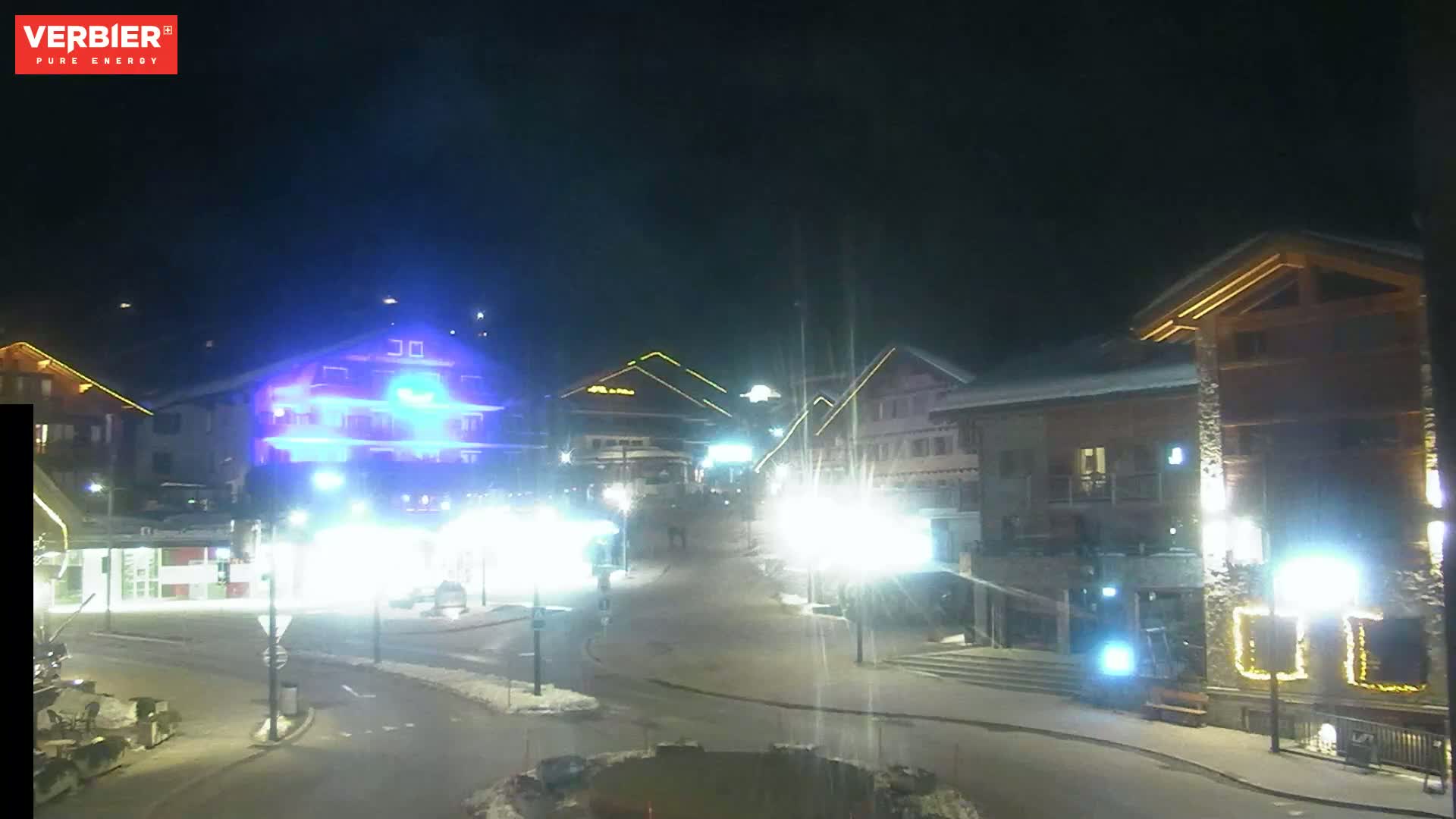 A nighttime view of a snow-covered town square with brightly lit buildings and streetlights.