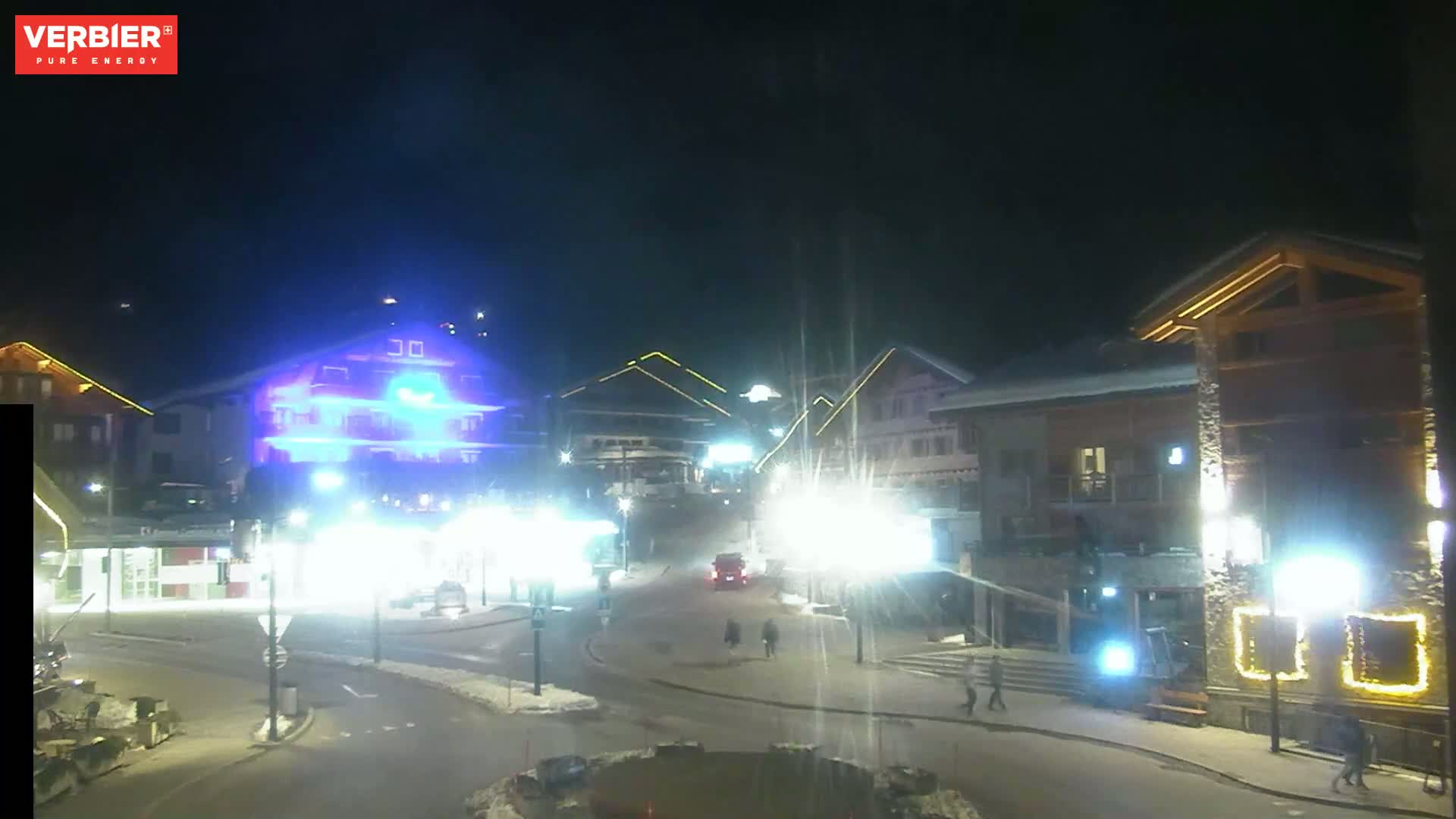A brightly lit town square at night, with several buildings and pedestrians visible under a clear, dark sky.
