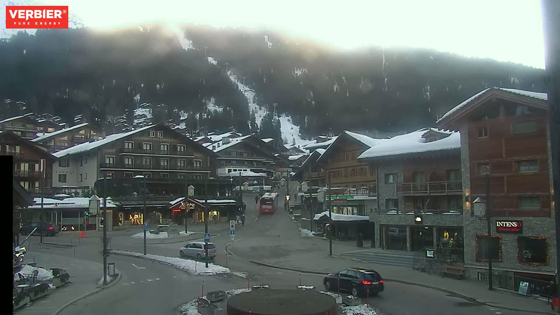 A snow-covered village nestled in a valley under a hazy, bright sky, with a few cars and a bus on the road.
