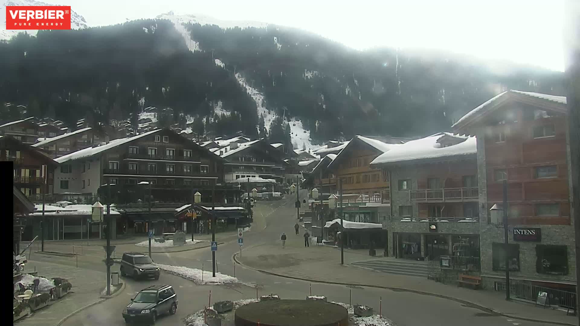 A snow-covered village nestled in a valley beneath a misty, snow-dusted mountain, with a few pedestrians and cars visible on the street.