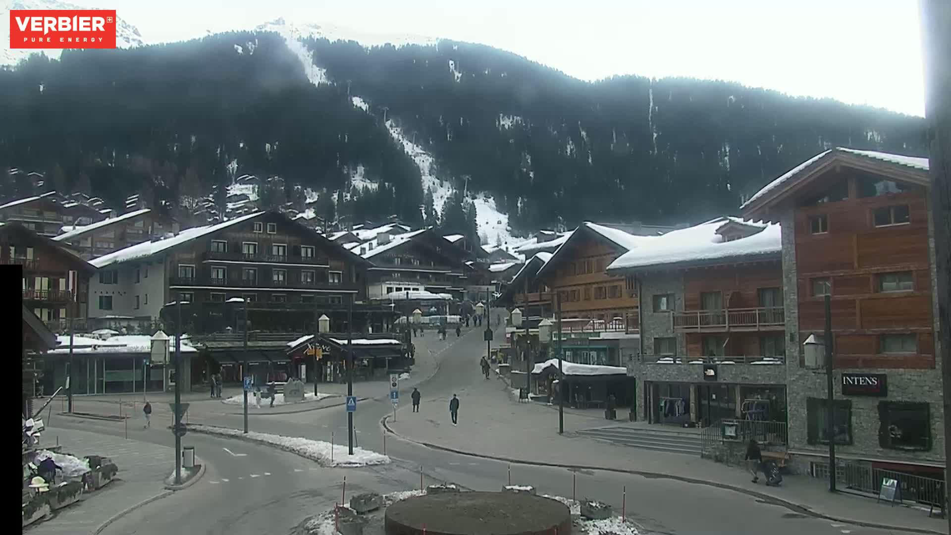 A snow-covered village nestled in a mountain valley on an overcast day, with a few people walking in a plaza.
