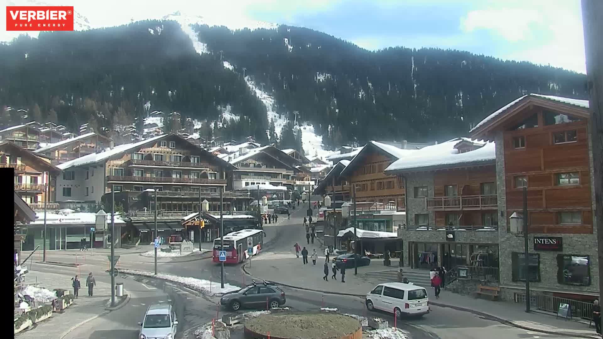 A snow-dusted village nestled in a mountain valley under a sunny sky, with pedestrians and vehicles on the street.