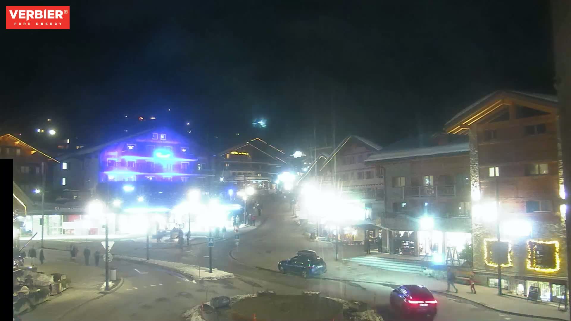 A nighttime scene of a snow-covered town square with several buildings lit up, cars parked, and a few pedestrians walking around.