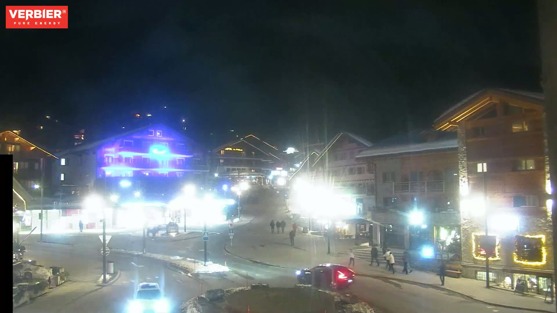A nighttime scene of a snow-dusted town square with brightly lit buildings and a few pedestrians, under a dark sky.