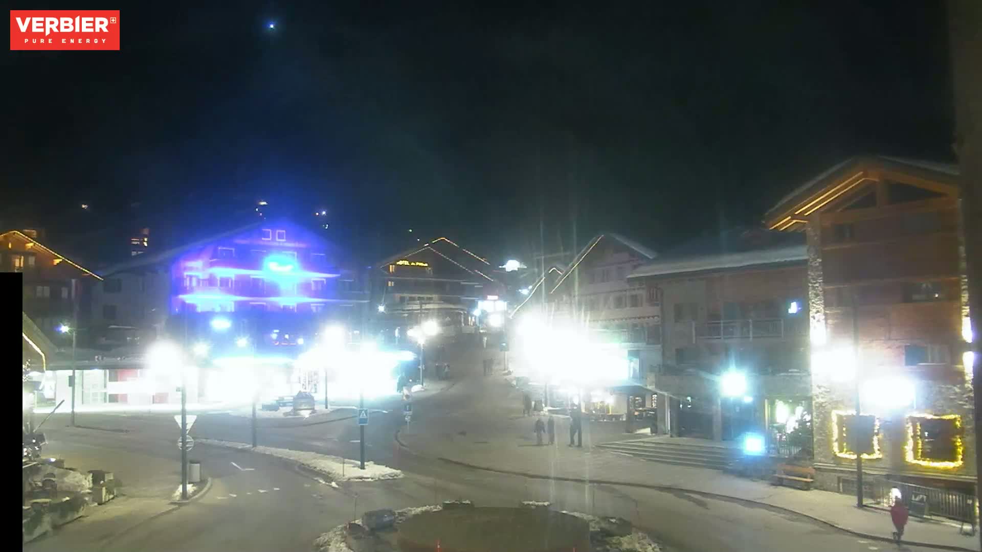 A nighttime view of a snow-covered town square with several brightly lit buildings and a few pedestrians.