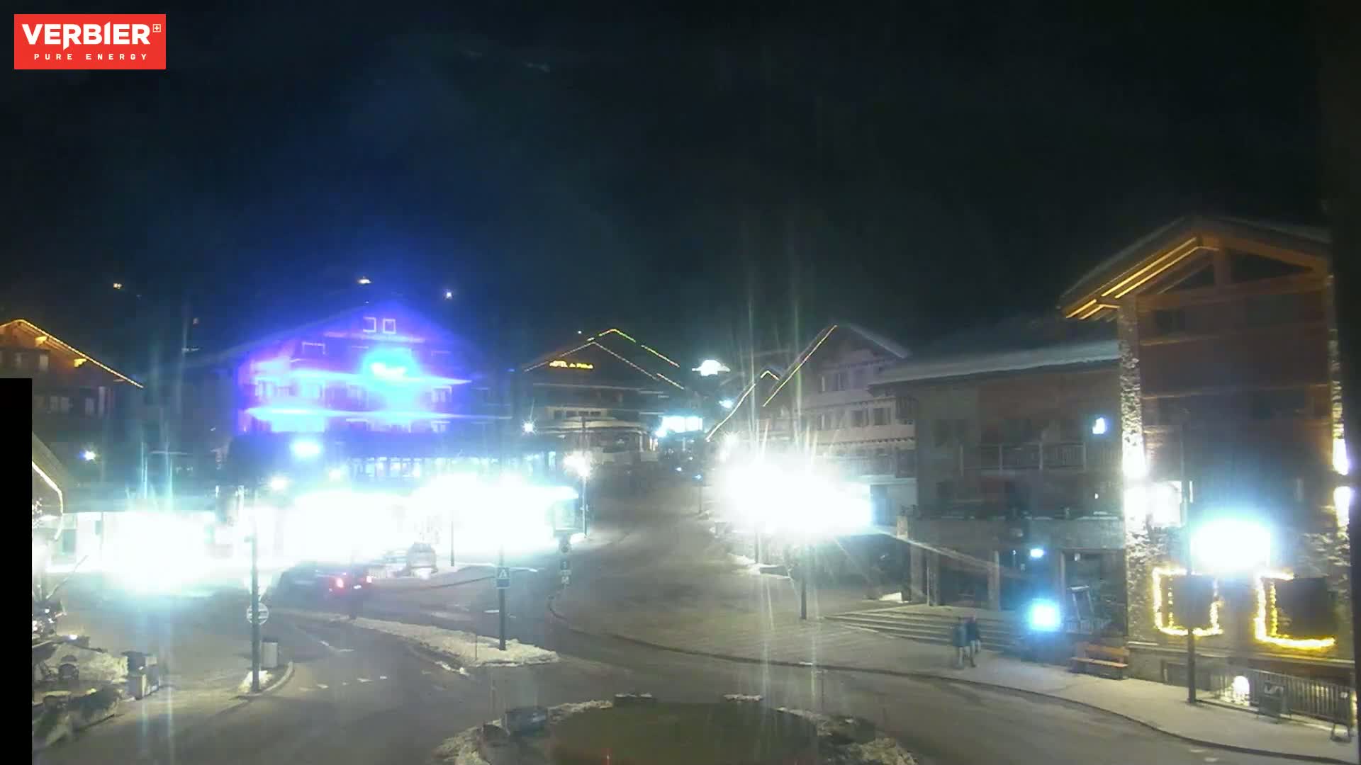 A nighttime view of a snow-covered town square with brightly lit buildings and streetlights.