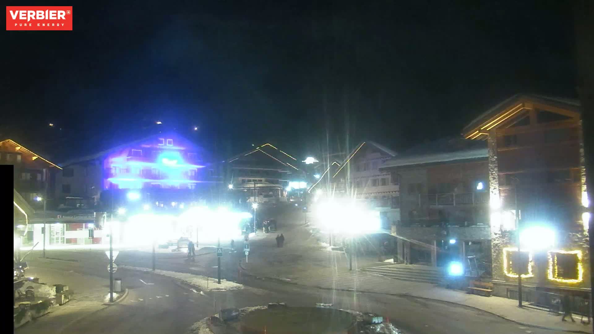 A nighttime view of a snow-covered town square with brightly lit buildings and a few pedestrians.
