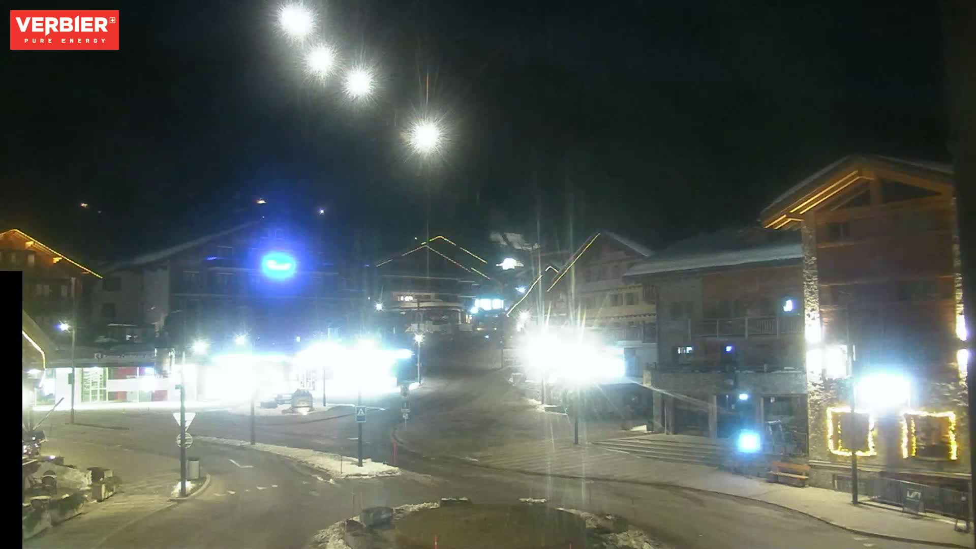 A nighttime view of a snow-covered town square with brightly lit buildings and streetlights.