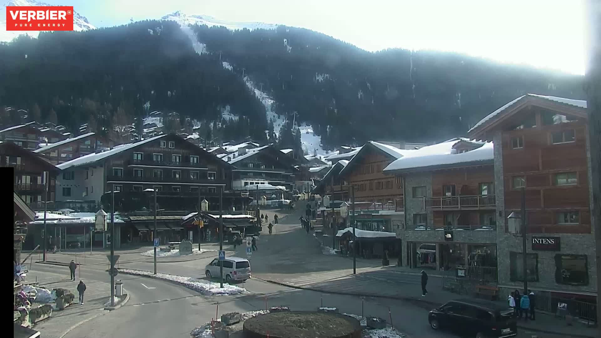 A sunny day in a snow-covered mountain village, with several buildings and a few cars and people visible in a town square.