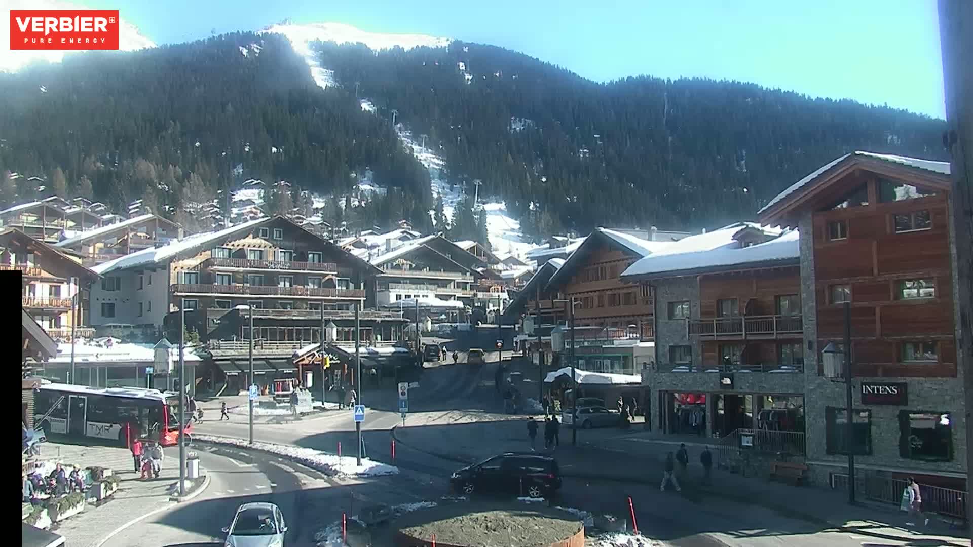 A sunny day in a snow-dusted alpine village with several buildings, a few cars, and people walking around, situated at the base of a snow-covered mountain with ski runs visible.