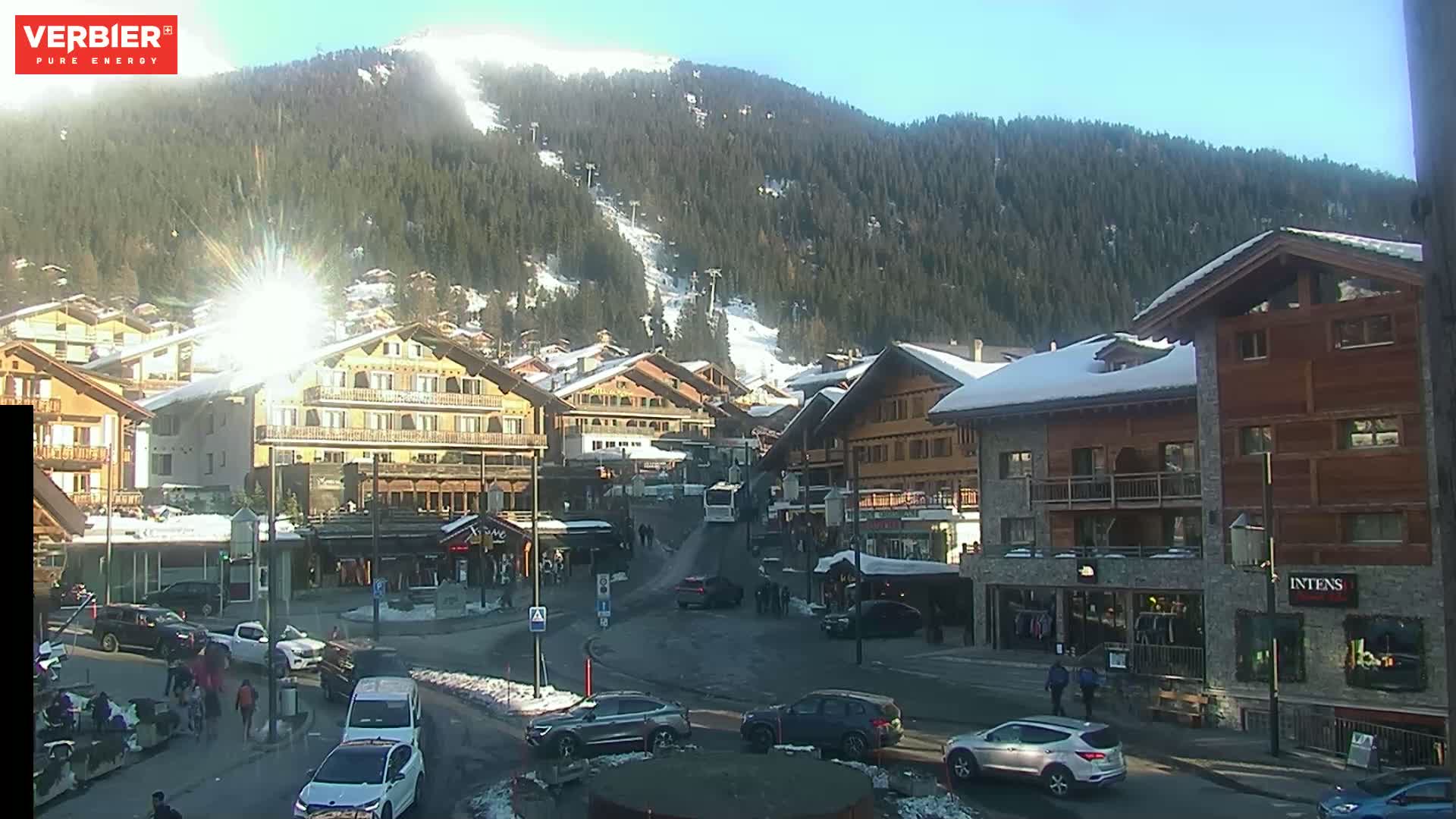 A sunny day in a snow-covered mountain village with several buildings, cars, and people around a small roundabout.