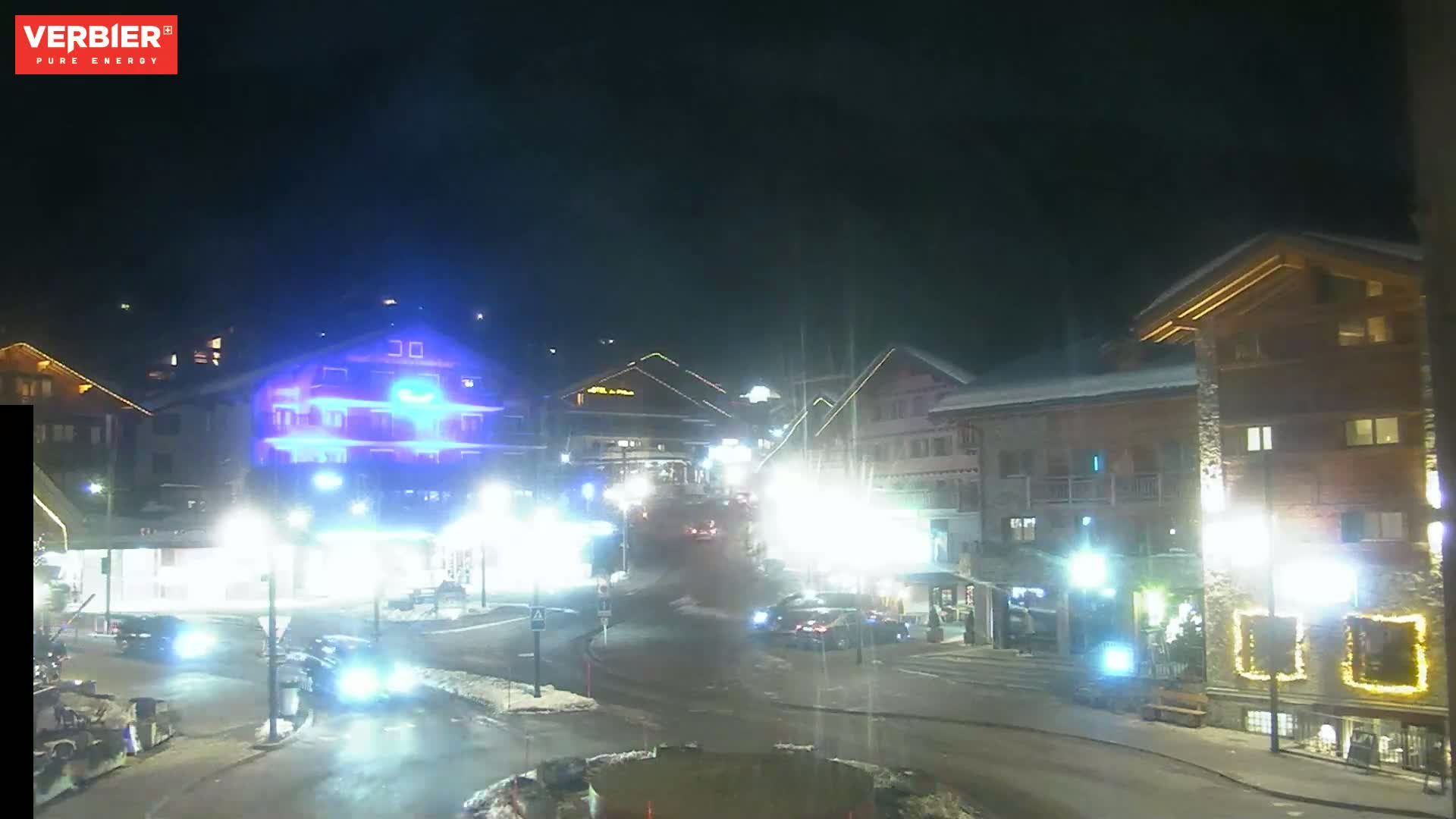A nighttime view of a town square with several buildings illuminated, vehicles on the road, and some snow on the ground.