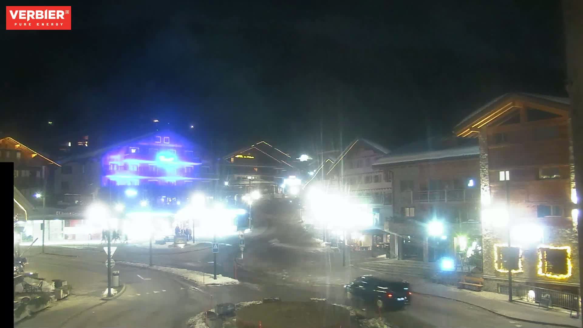 A nighttime view of a snow-dusted town square with brightly lit buildings and cars driving on the road.
