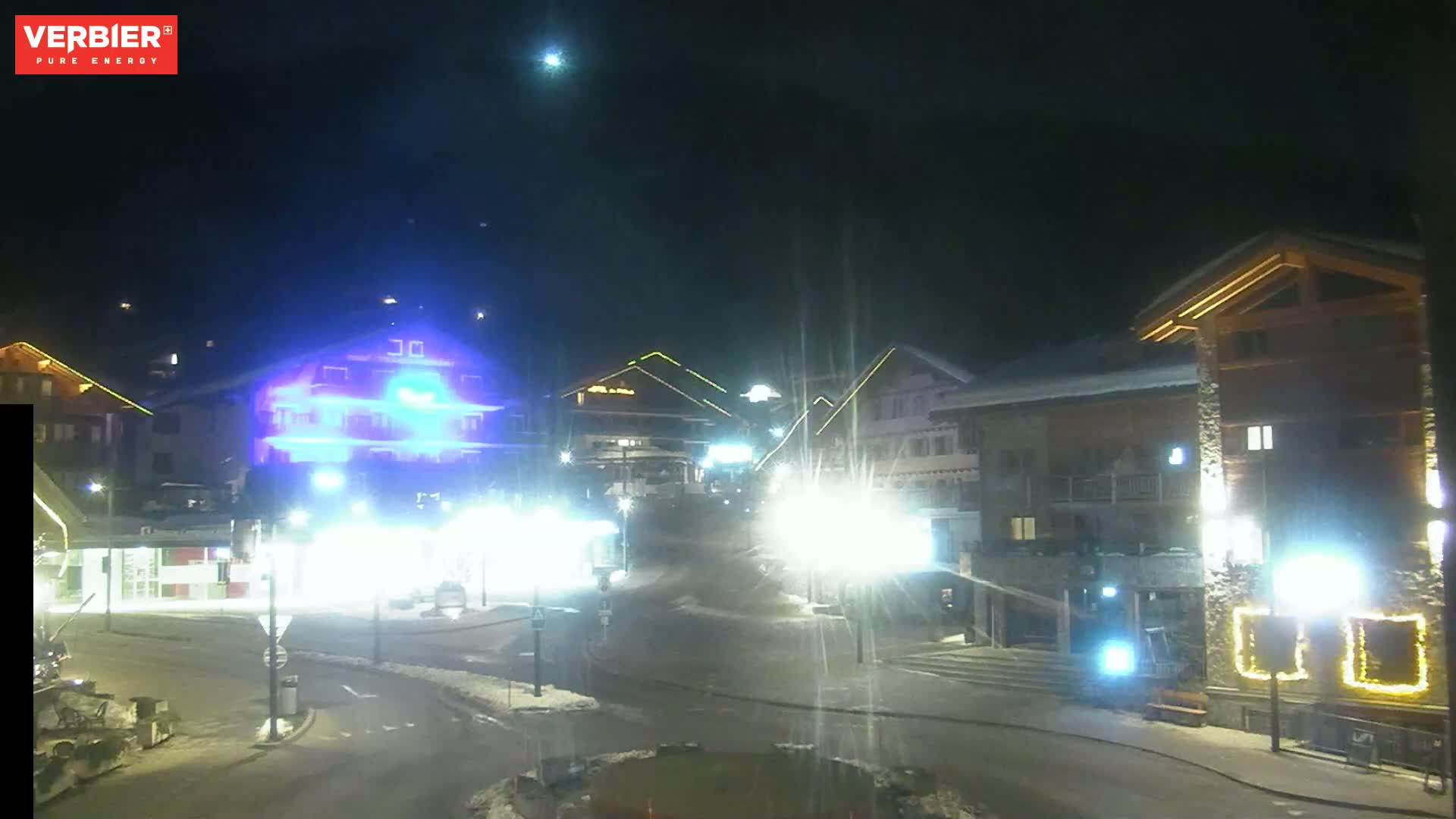 A nighttime scene of a snow-covered town square with brightly lit buildings and streetlights.