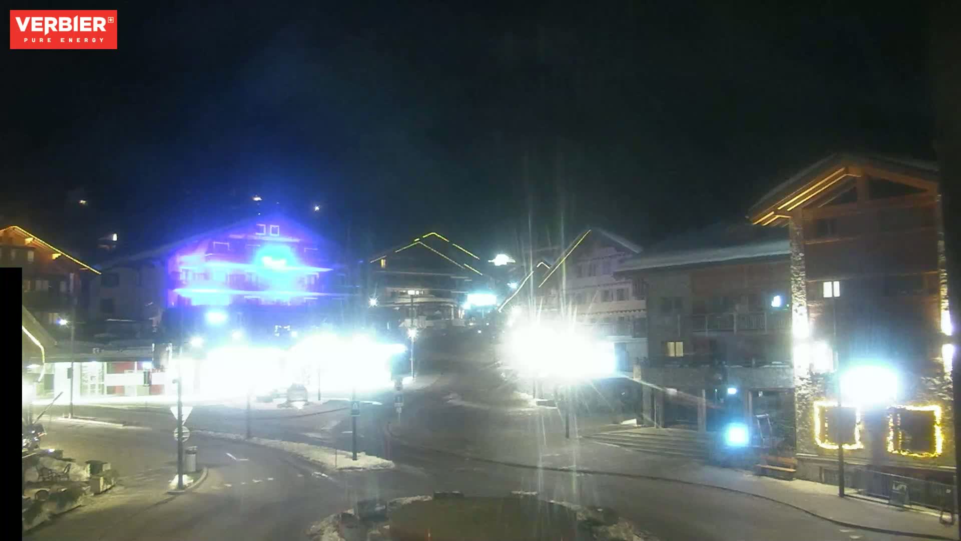 A nighttime view of a snow-covered village square with brightly lit buildings and streetlights.