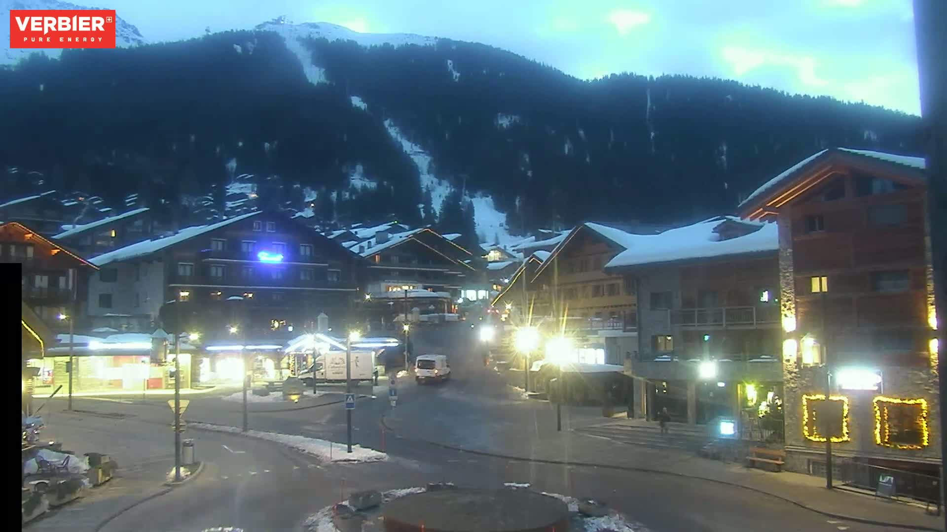 A snow-covered mountain village at dusk, with illuminated buildings and a mostly clear sky.
