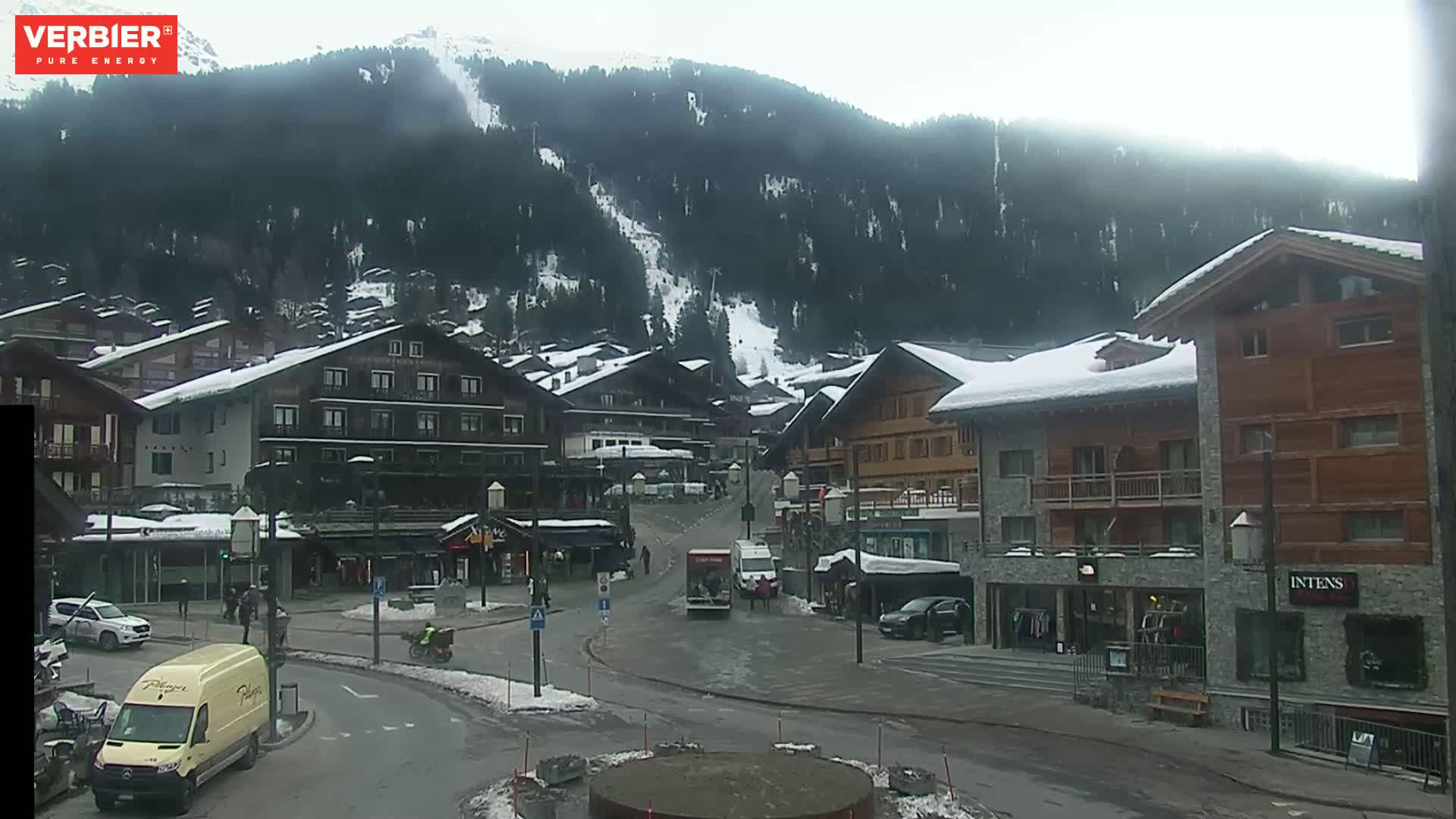 A snow-covered village nestled in a mountain valley on an overcast day, with several vehicles parked and moving in the streets.
