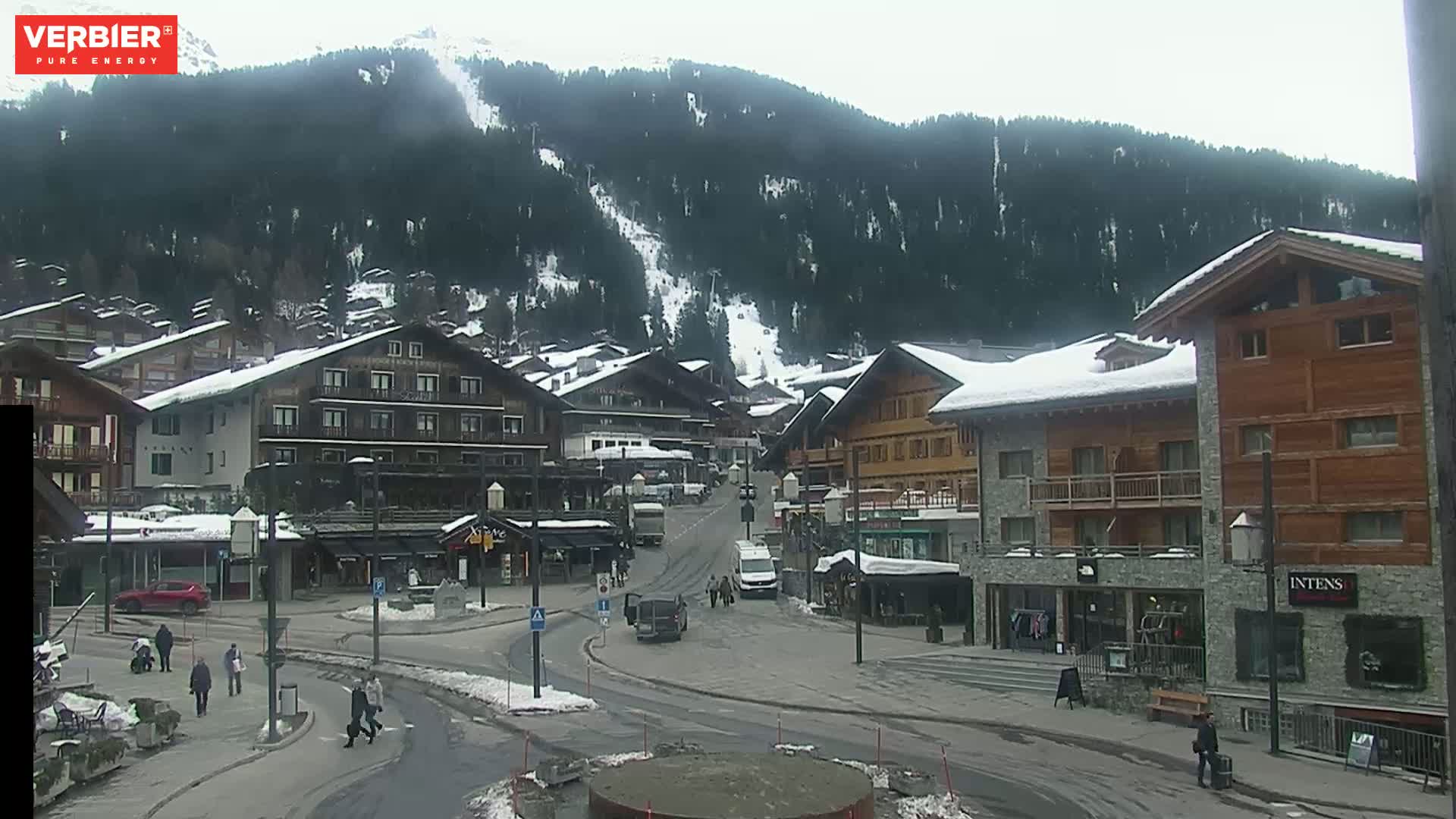 A snowy village square with several buildings, some cars, and a few pedestrians is nestled at the base of a snow-dusted mountain range on an overcast day.