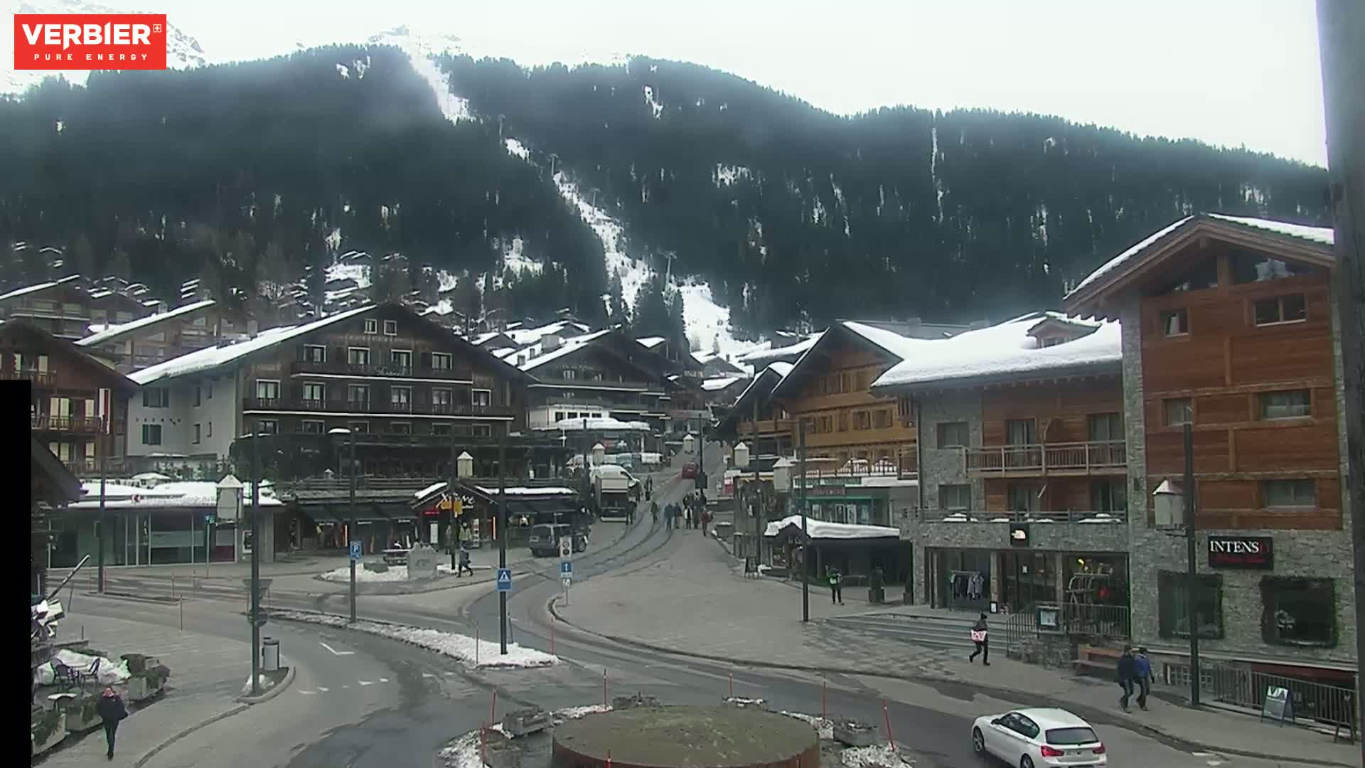 A snow-covered village nestled in a valley surrounded by a snow-dusted mountain range under an overcast sky.