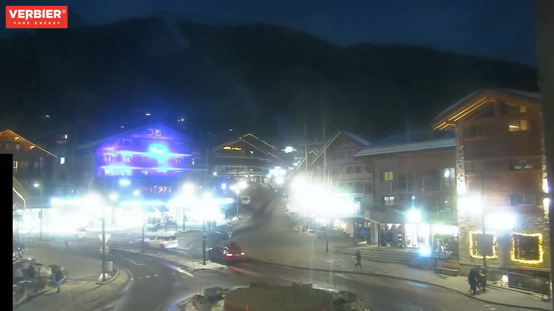 A nighttime view of a snow-dusted village nestled at the base of a dark mountain, illuminated by streetlights and building lights.