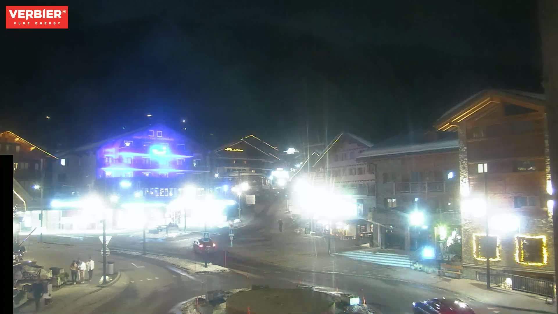 A nighttime view of a snow-covered town square with illuminated buildings and a few cars.