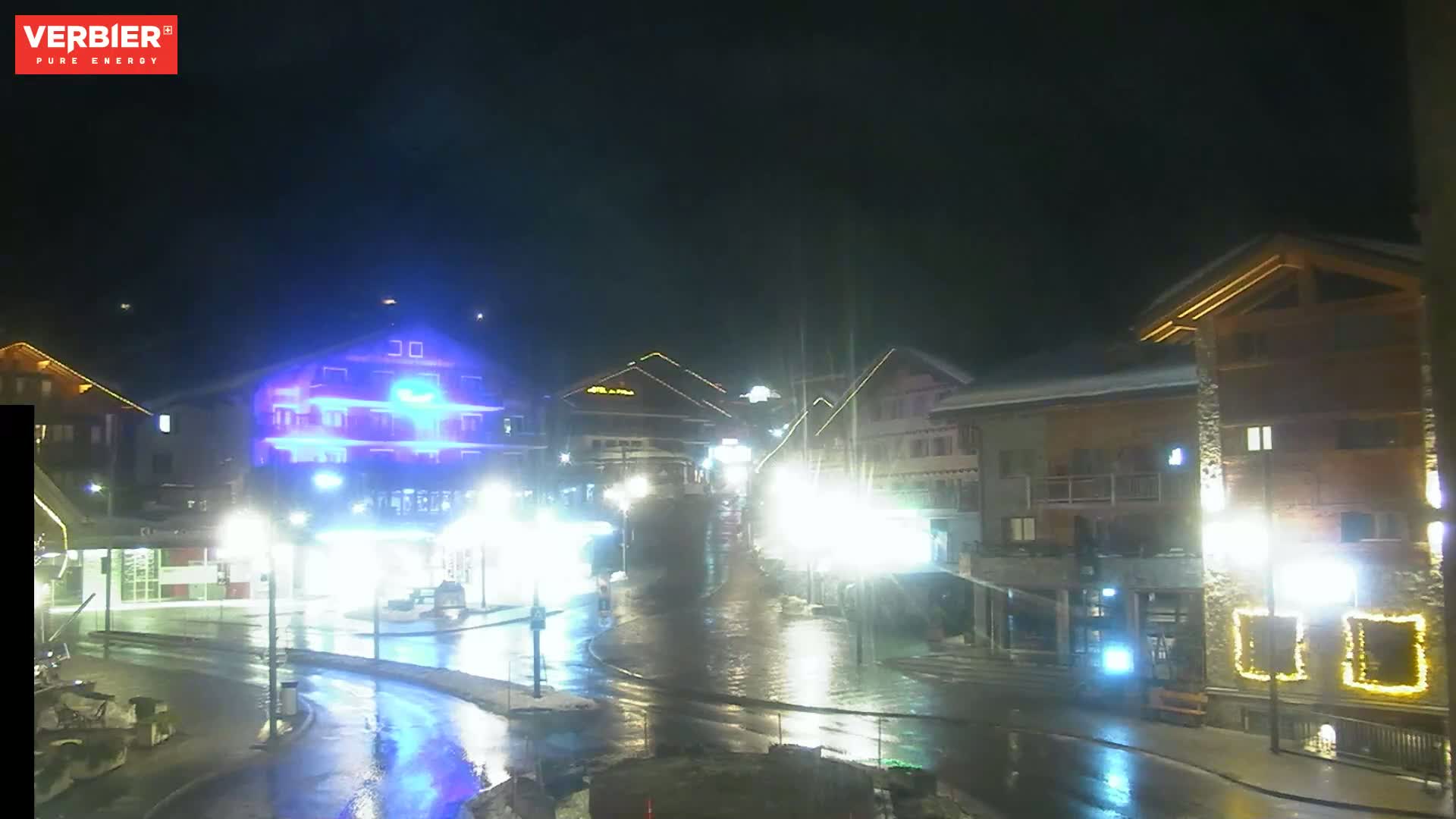 A wet, nighttime street scene shows brightly lit buildings, some with illuminated signs, in a town or village setting.