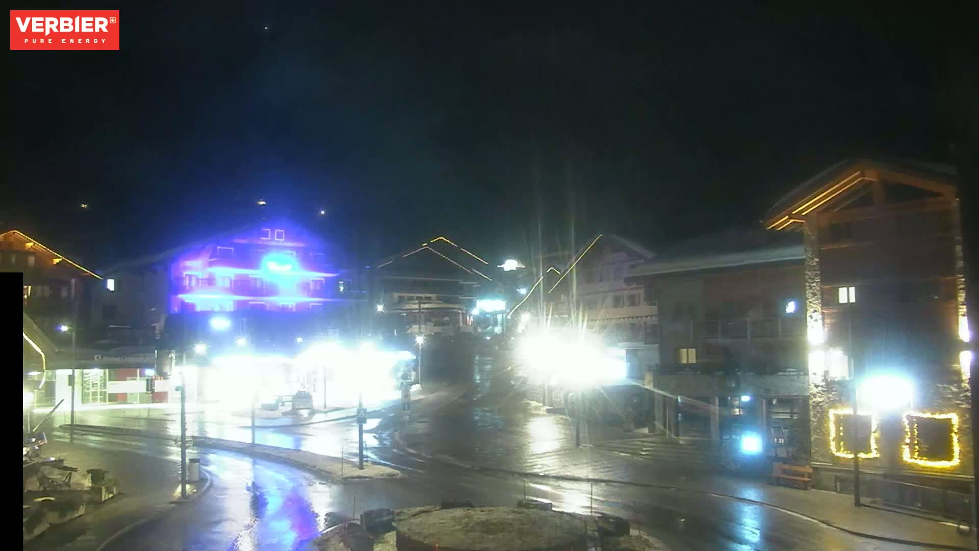 A wet, nighttime street scene shows several illuminated buildings and streetlights in a town square.