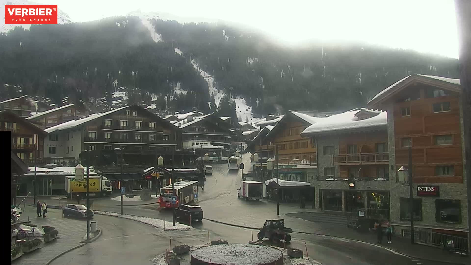 A snow-covered mountain village street scene on an overcast day, with several buildings, vehicles, and pedestrians visible.