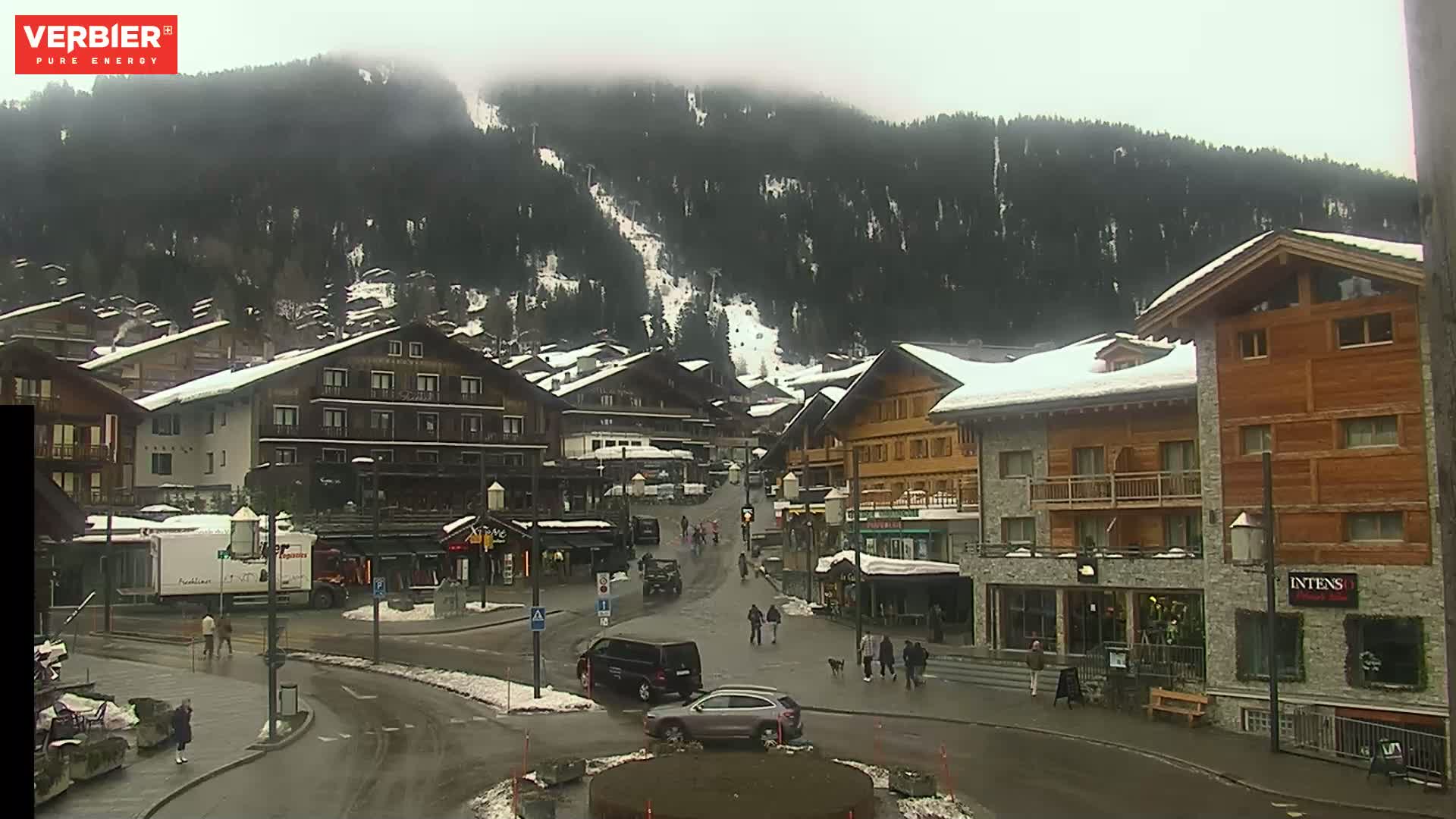 A mountain village street scene on an overcast day, with snow-dusted roofs and a few pedestrians walking amid several buildings.