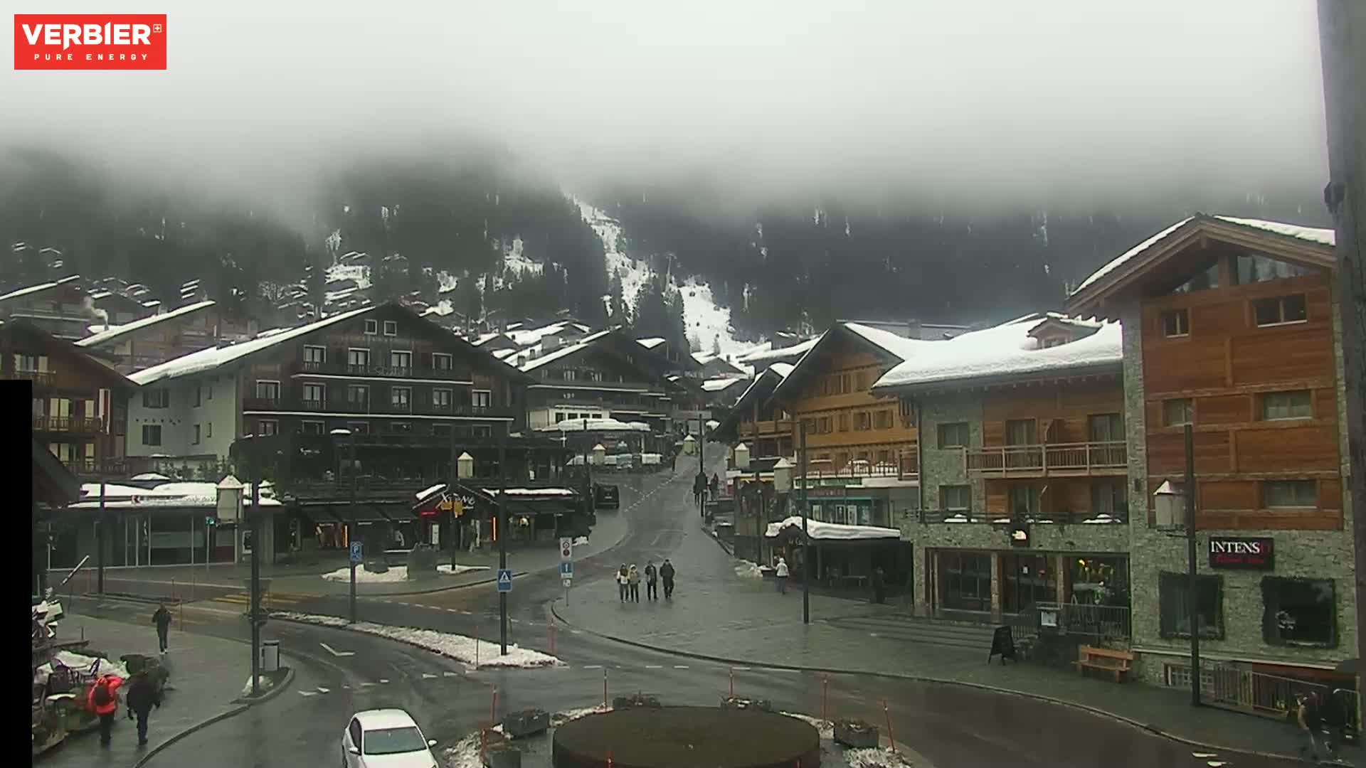 A snow-covered village nestled in a foggy mountainside, with several people walking on a wet street.