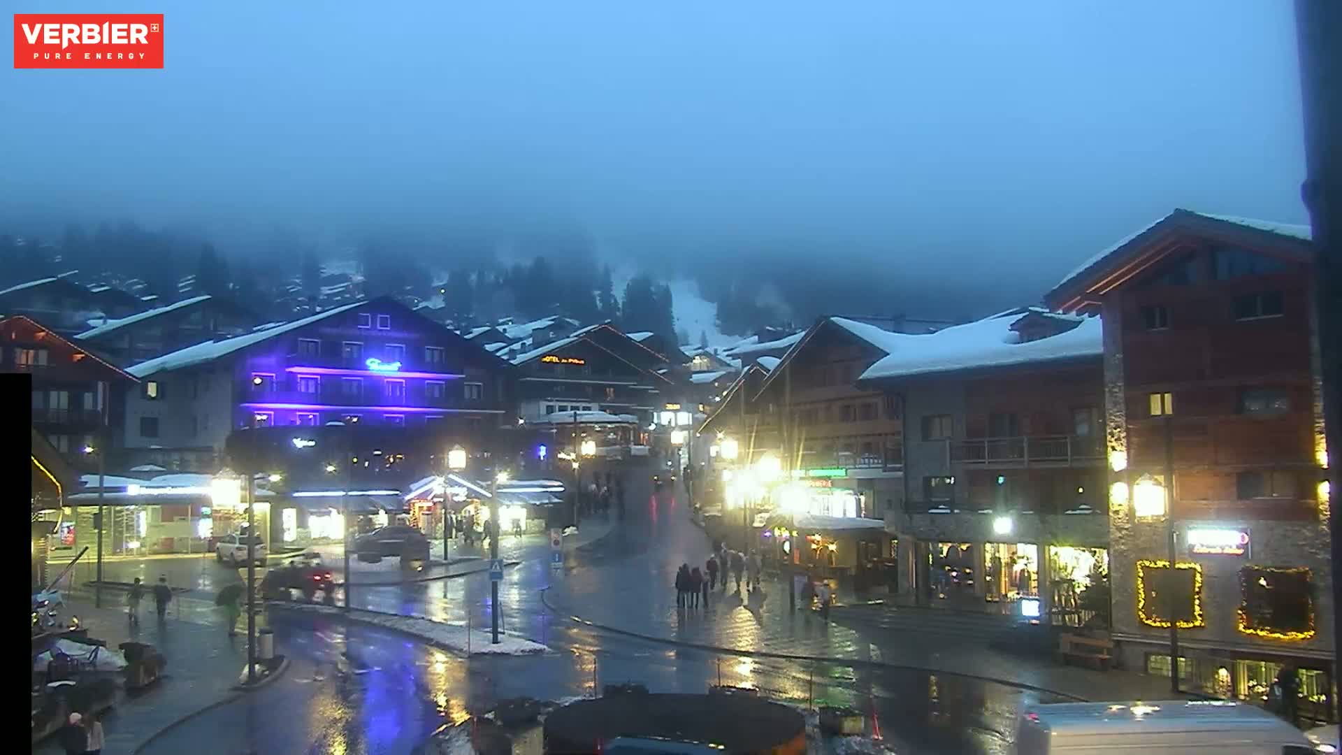A wet, dimly lit village square is surrounded by snow-covered buildings under a foggy, twilight sky.