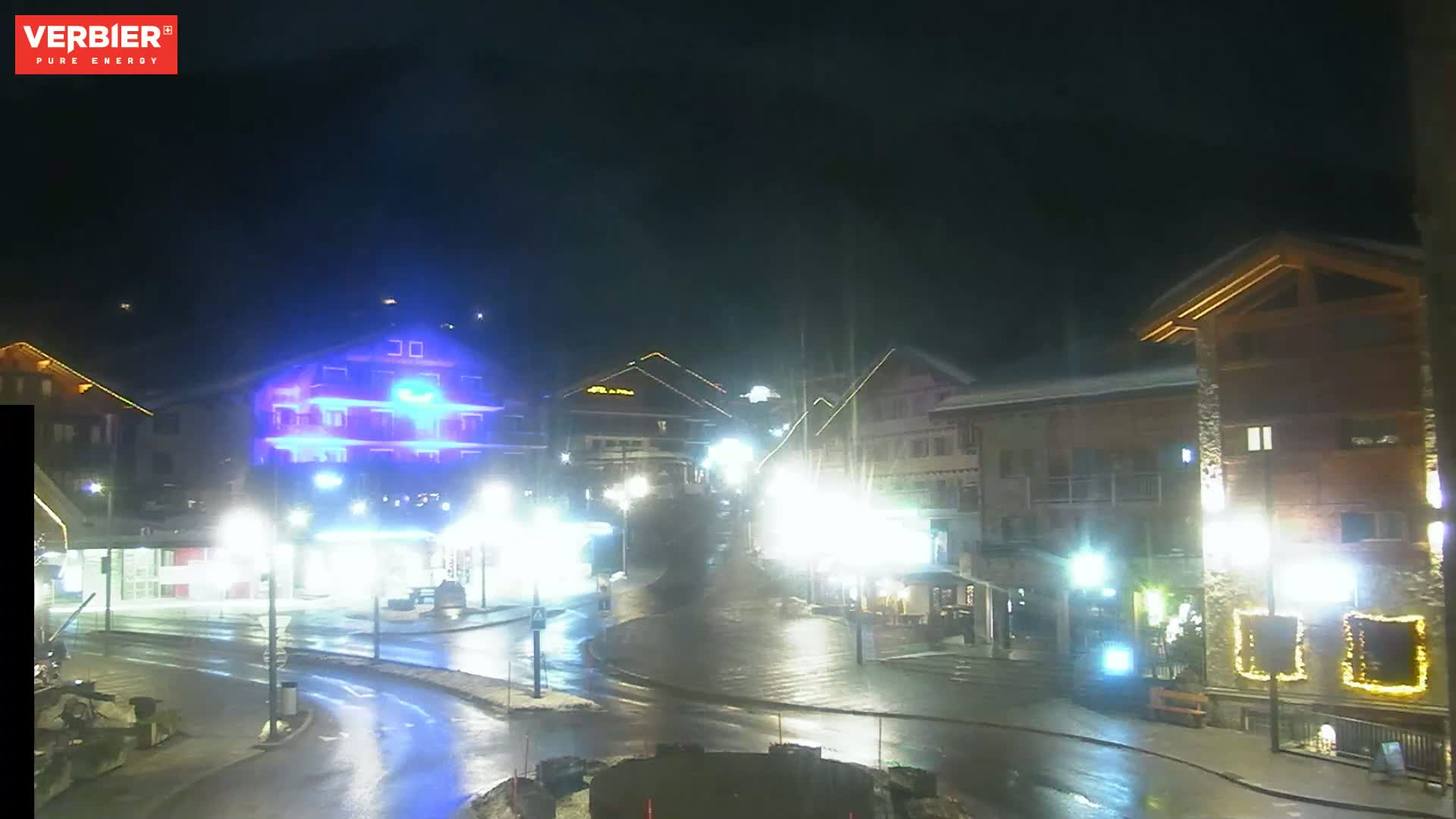 A nighttime view of a town square with several illuminated buildings, wet streets, and minimal traffic.