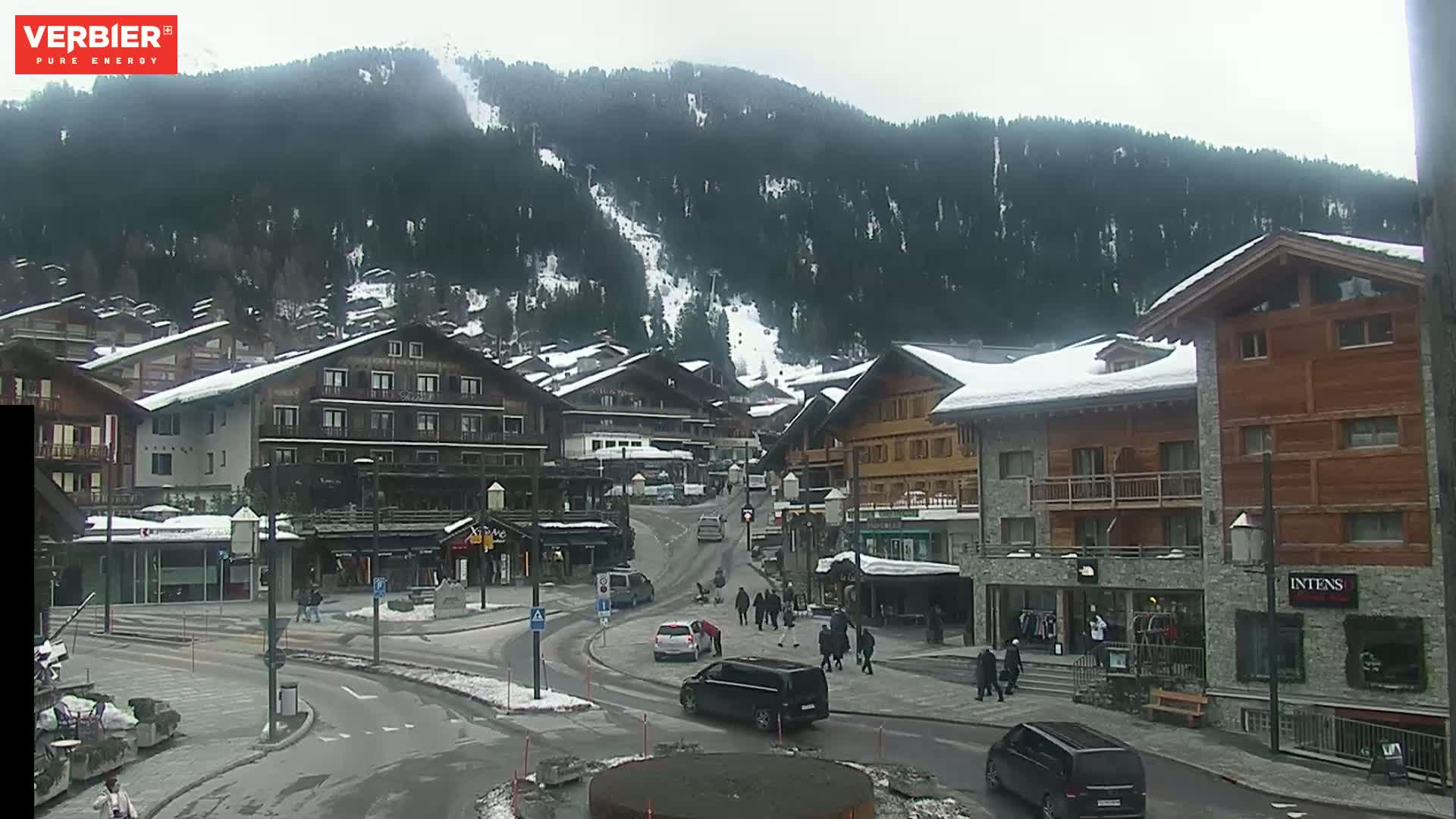 A snow-covered village nestled in a mountainous region on an overcast day, with cars parked along the roads and pedestrians walking near buildings.