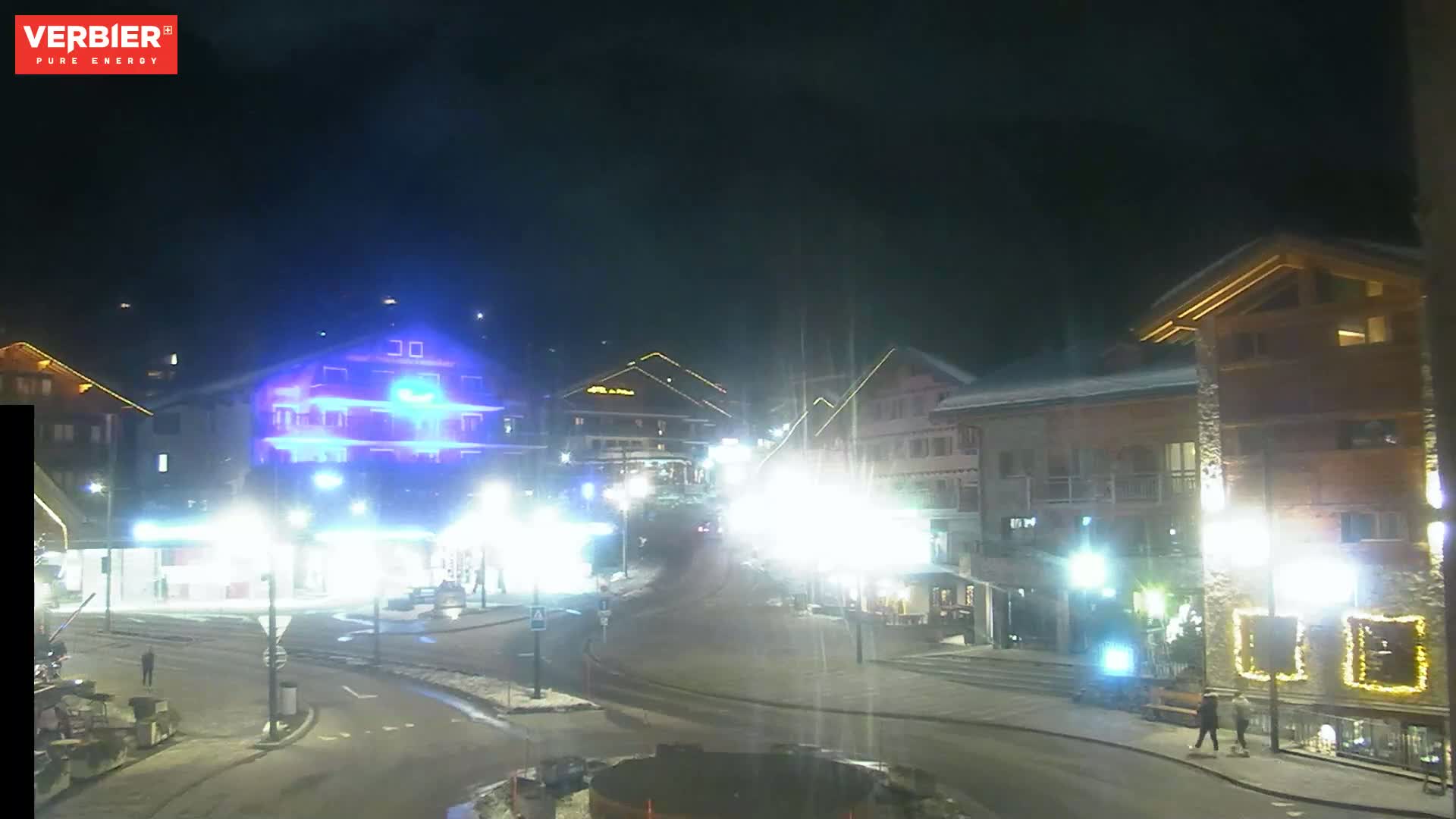 A nighttime view of a town square with several brightly lit buildings, and some pedestrians, under a dark, clear sky.