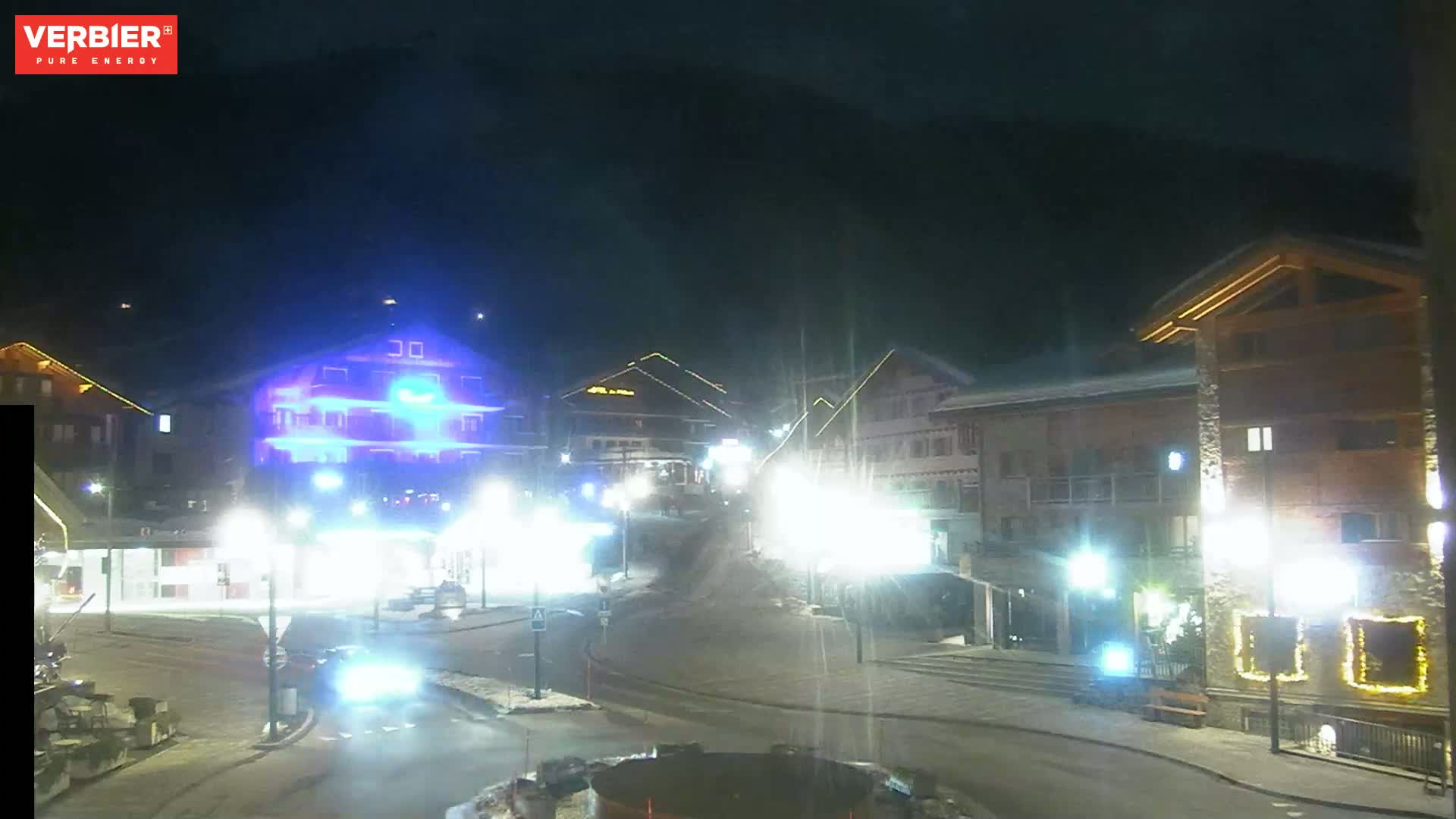 A nighttime view of a snow-covered town square with brightly lit buildings and streetlights, under a dark sky.