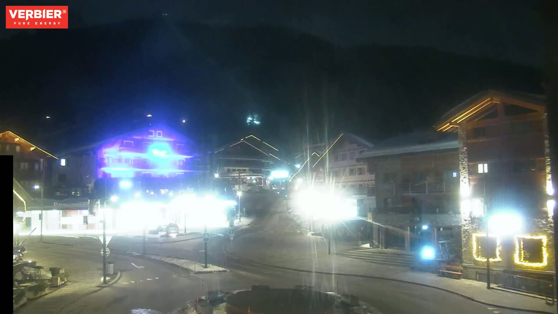 A nighttime, snow-covered street scene in a village, illuminated by streetlights and building lights against a dark mountain backdrop.