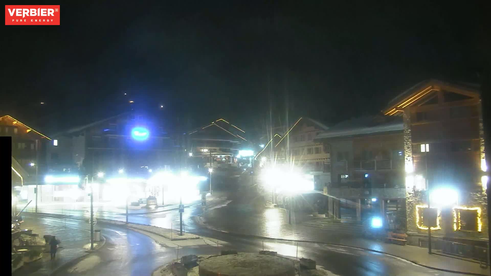 A wet, nighttime scene shows brightly lit buildings and a street with minimal traffic.