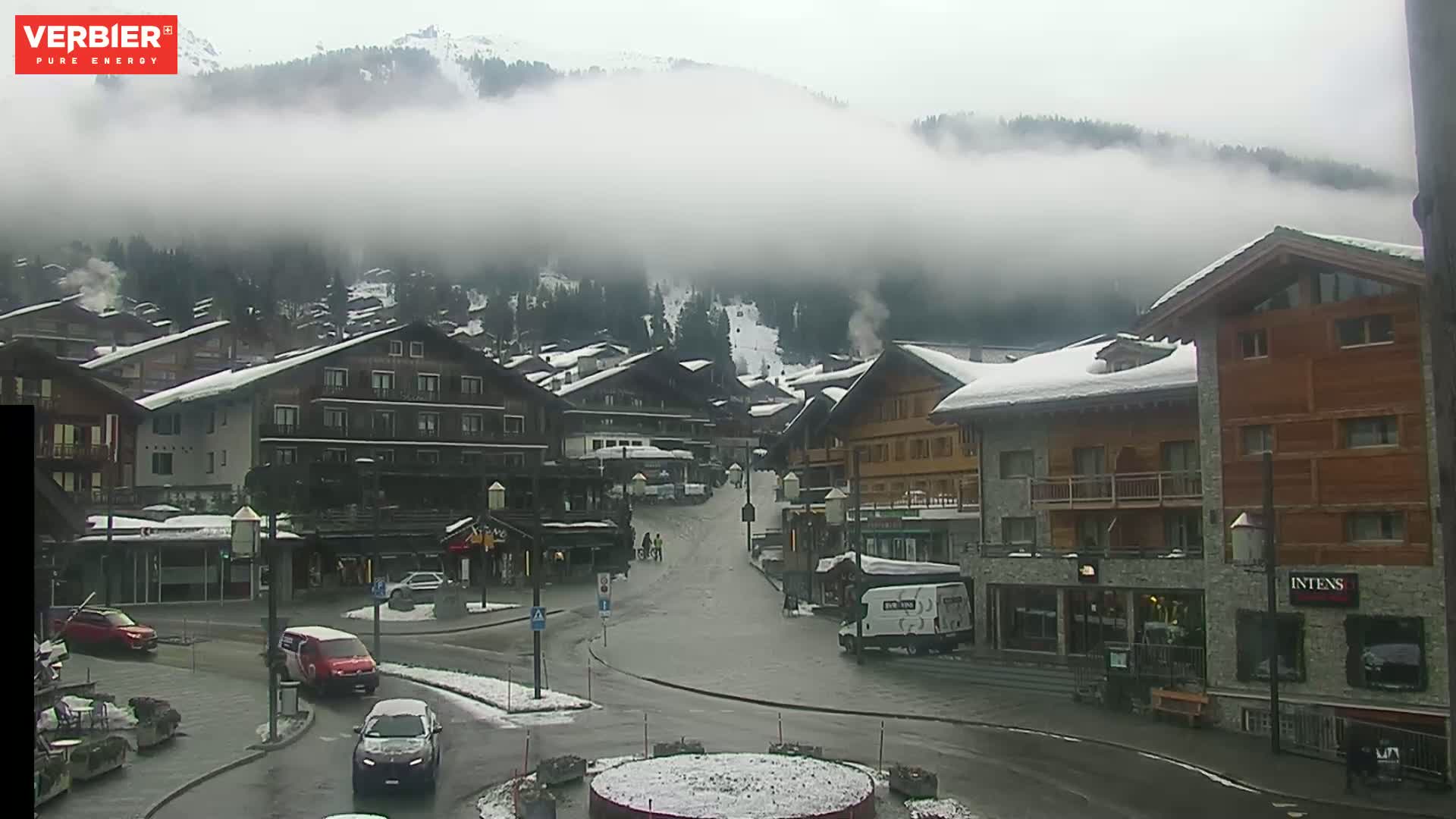 A snow-dusted village in a valley is shrouded in fog, with a few cars visible on the wet streets.