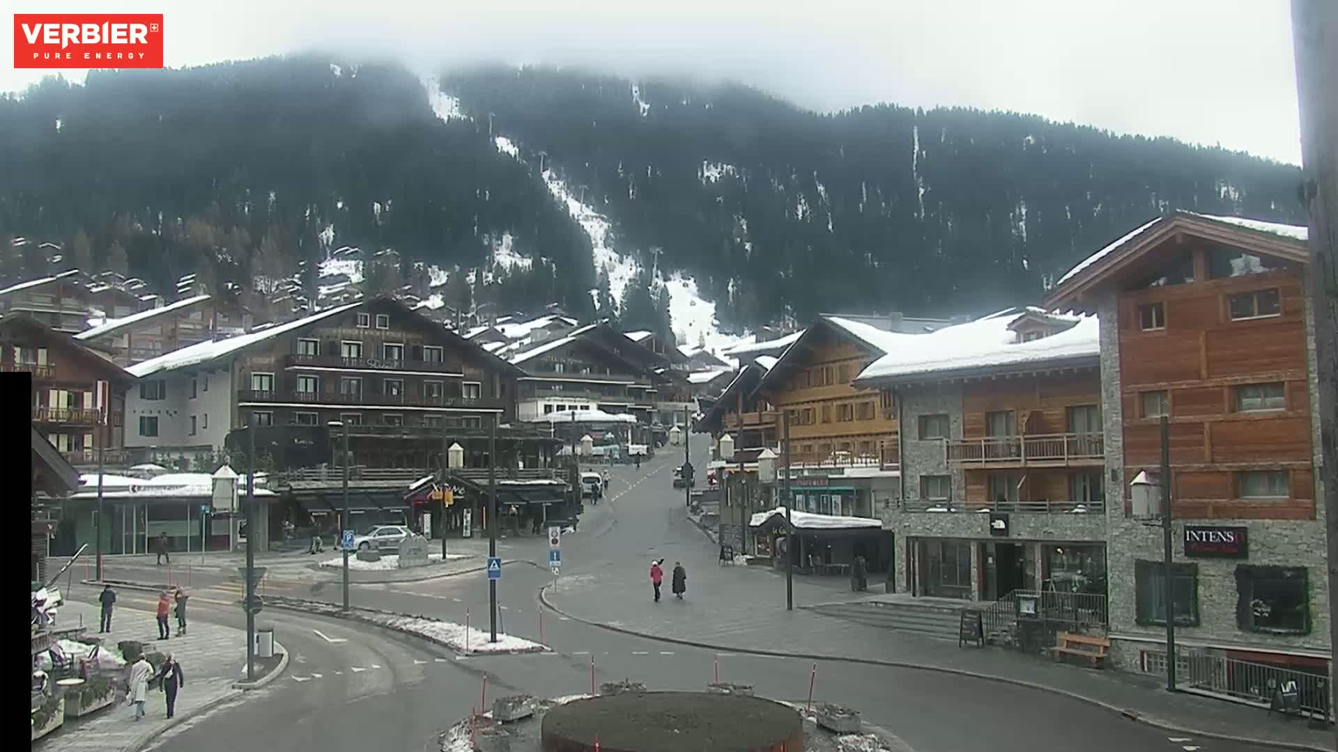 A snow-dusted village nestled in a valley beneath a cloudy sky, with a few pedestrians walking along the streets.