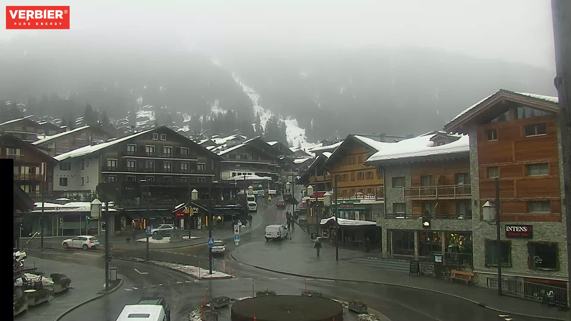 A snow-dusted village in a foggy mountainous area, with several cars parked and driving on wet roads.