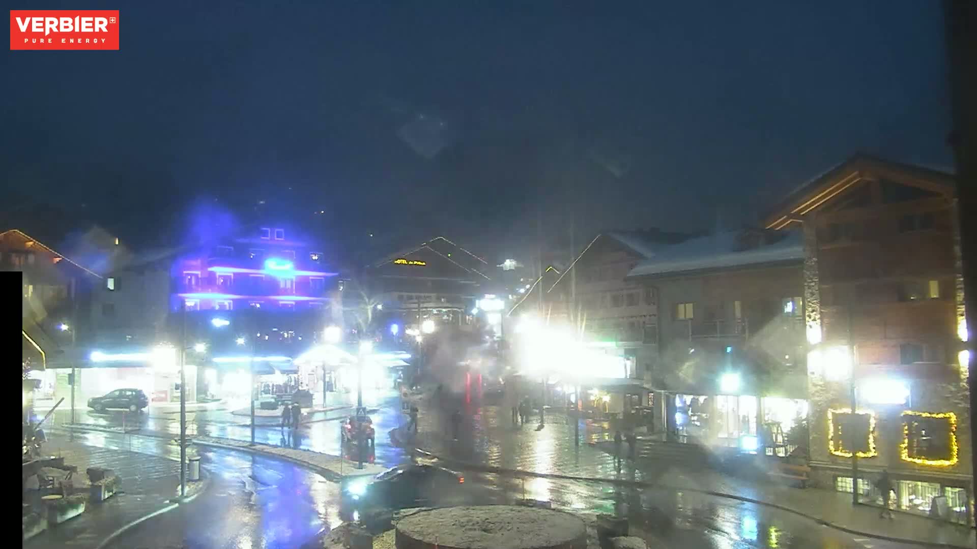 A wet, brightly lit town square at night, with several buildings and some pedestrians visible under a dark sky.