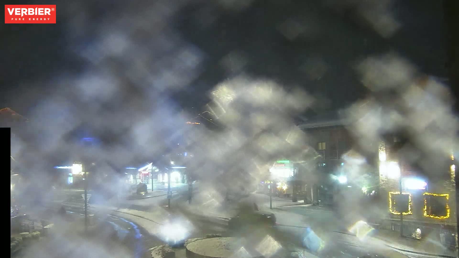 A nighttime scene of a snow-covered town square obscured by heavy fog or steam, with buildings and streetlights faintly visible.